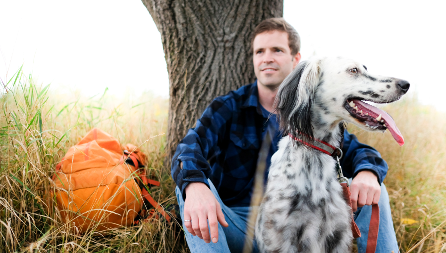 Mann sitter på gresset foran et tre med sin oransje ryggsekk med hunden sin foran seg