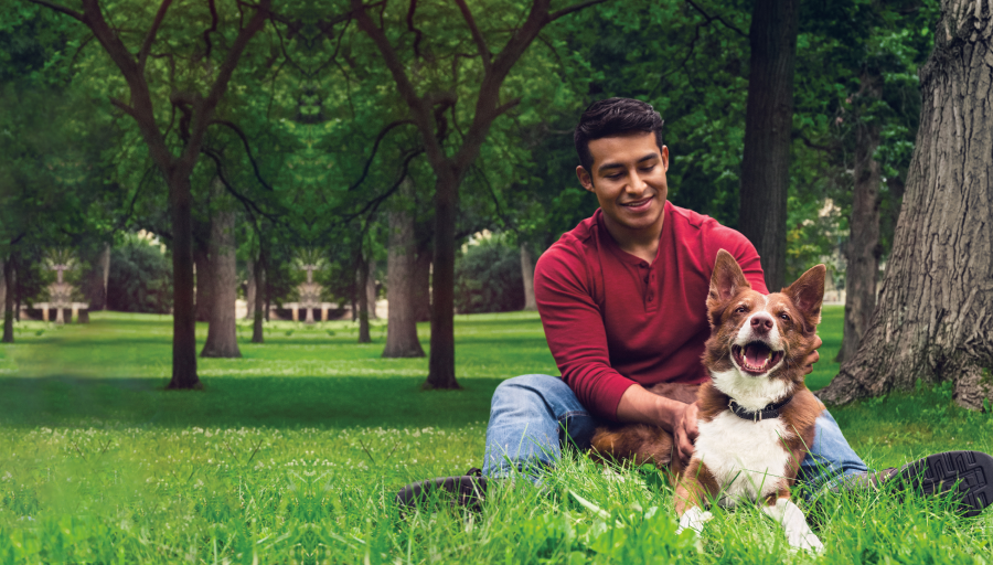 Mann sitter på gresset med trær rundt seg med hunden liggende foran seg