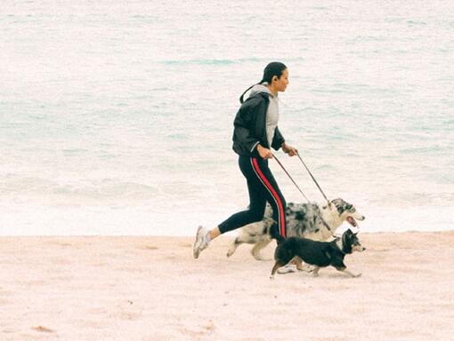 En kvinne med to hunder løper langs stranden